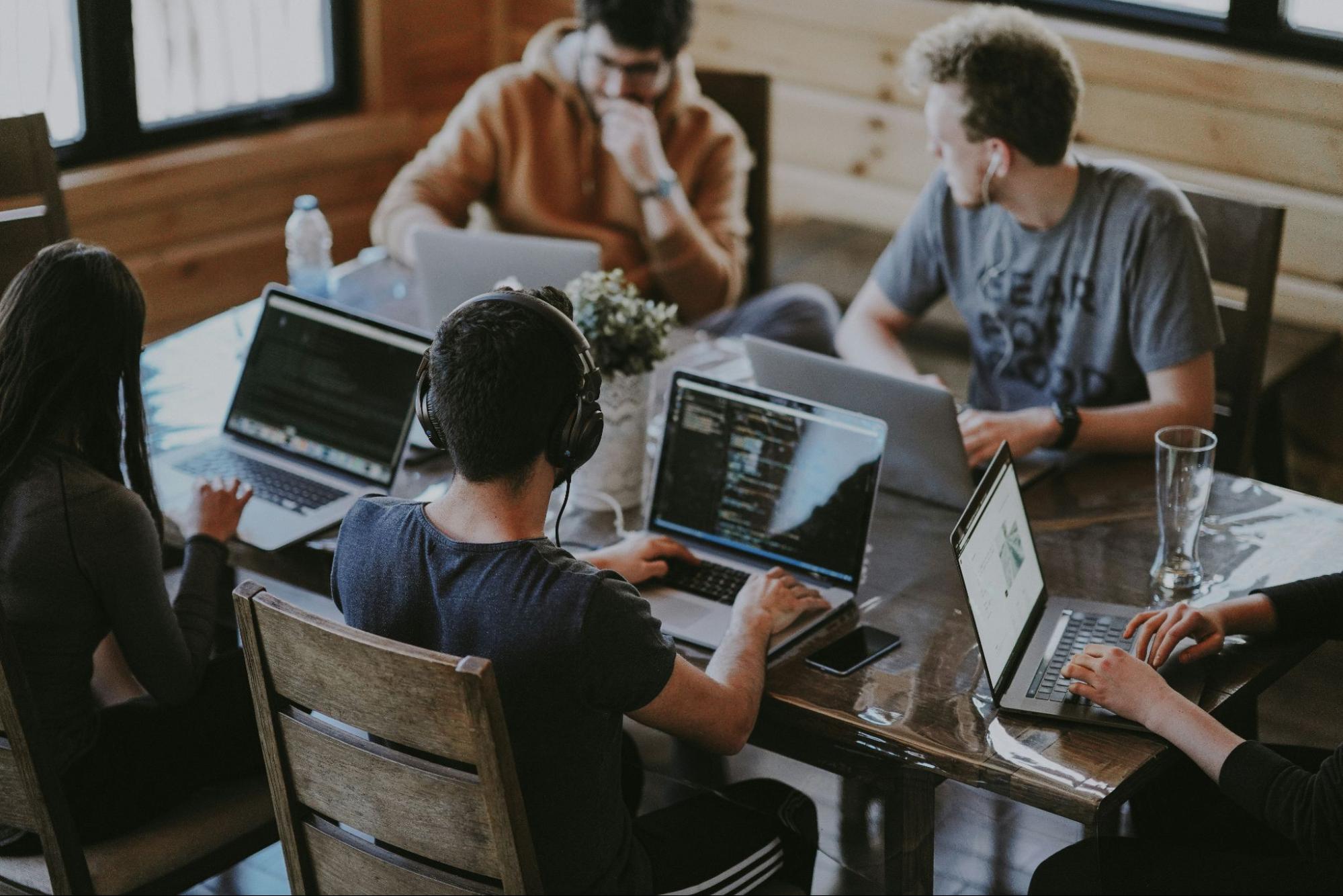 A group of young professionals working together at a table with laptops. One person wears headphones, while others engage in coding and discussions, creating a collaborative and productive workspace environment. (source: Unsplash)