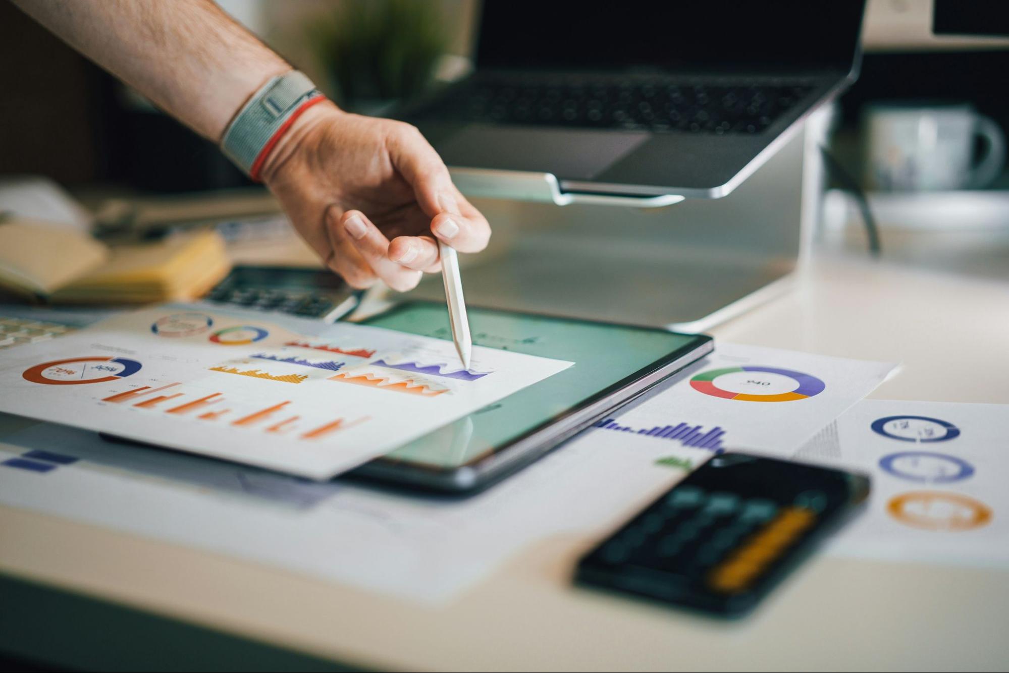 Close-up of a person's hand using a stylus on a tablet, with colorful analytics charts and graphs on paper in the foreground, and a laptop in the background. Represents data analysis and digital tools in a professional workspace. (source: Unsplash)